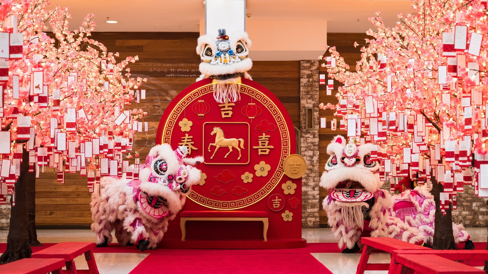 Traditional Chinese lion dance performance with red backdrop and festive decorations in a mall setting.